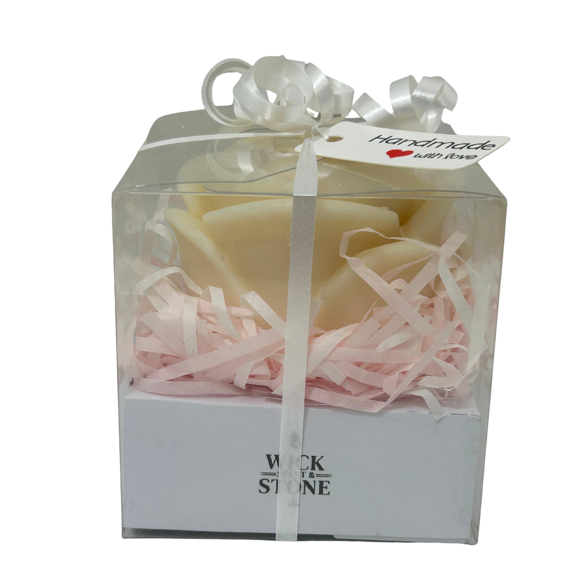 Candle in a clear box with pink shredded paper and a white ribbon, branded 'Wick & Stone'.